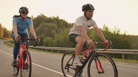 Un-Hombre-Y-Una-Mujer-En-Bicicleta-Recorren-Juntos-La-Carretera-Al-Atardecer-En-Cámara-Lenta.-La-Pareja-Viaja-En-Bicicleta.-Cascos-De-Ciclismo-Deportivos.